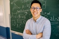Smiling teacher with glasses stands confidently before a math-covered blackboard.
